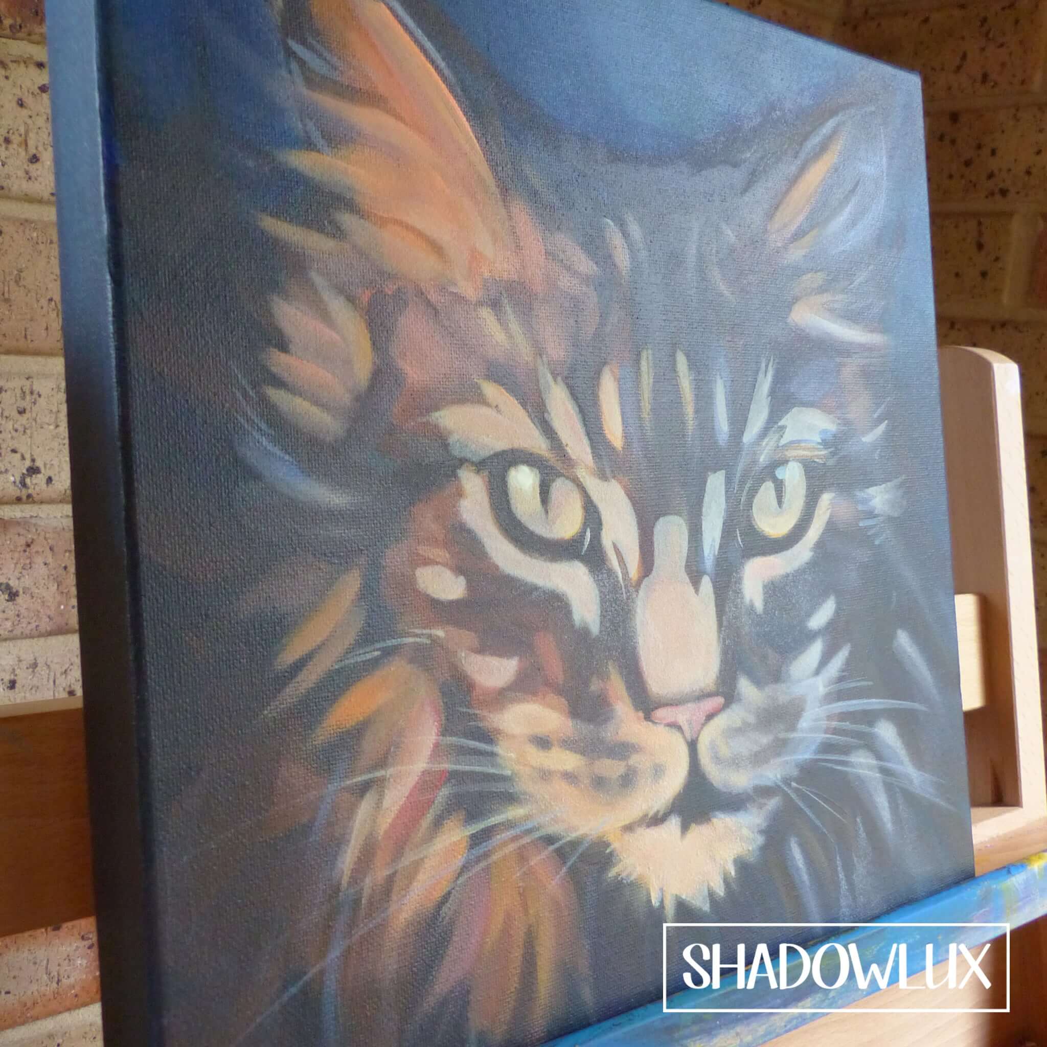 Painting on canvas of a Maine Coon cat with long fur and tabby markings, shown in natural daylight on an artists easel